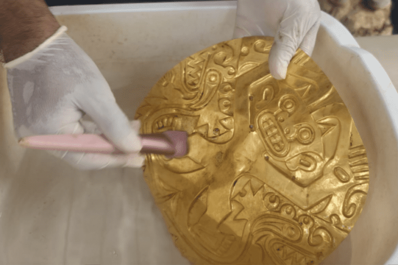 Κοντινό πλάνο ενός συντηρητή με λευκά γάντια που καθαρίζει προσεκτικά ένα μεγάλο, ανάγλυφο χρυσό θώρακα (pectoral) μέσα σε μια λεκάνη με νερό, χρησιμοποιώντας ένα μαλακό πινέλο.