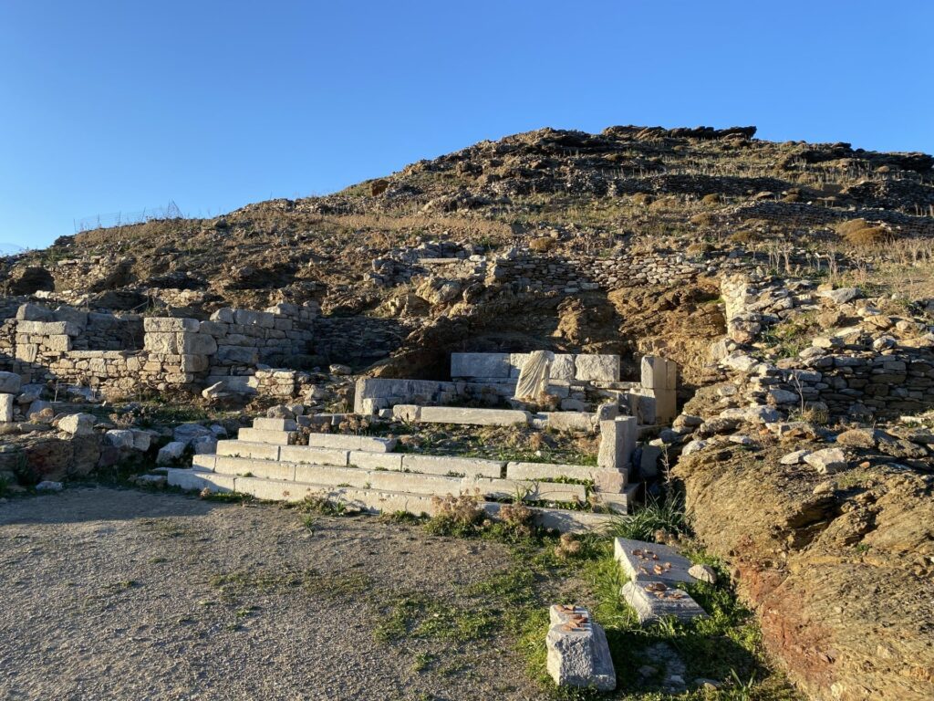 Μια ευρεία λήψη εξωτερικού χώρου αρχαιολογικών ερειπίων σε μια βραχώδη, ηλιόλουστη πλαγιά λόφου στα Κατάπολα της Αμοργού, με θέα σε ένα λιμάνι και το Αιγαίο Πέλαγος κάτω, κάτω από καθαρό μπλε ουρανό. Τα ερείπια περιλαμβάνουν πέτρινα θεμέλια, τοίχους και μια ανακατασκευασμένη σκάλα.