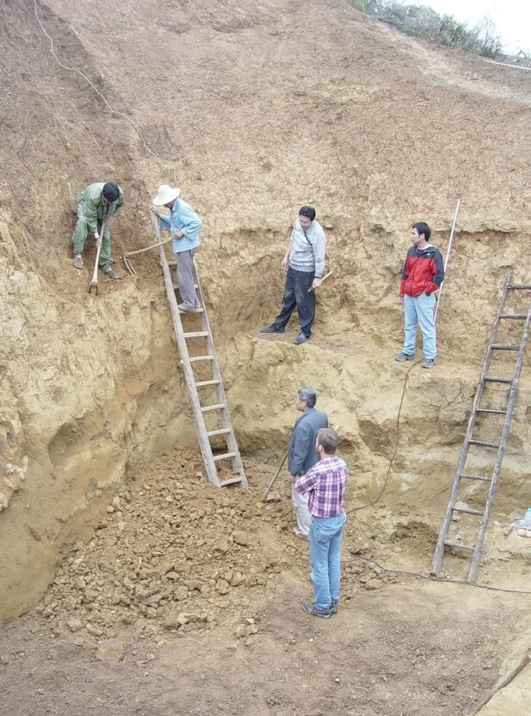 Ομάδα αρχαιολόγων και εργατών πραγματοποιεί ανασκαφή σε ένα βαθύ σκάμμα με χωμάτινα τοιχώματα. Χρησιμοποιούν ξύλινες σκάλες και εργαλεία χειρός, ενώ μερικοί επιβλέπουν τις εργασίες στο εσωτερικό της ανασκαφής.