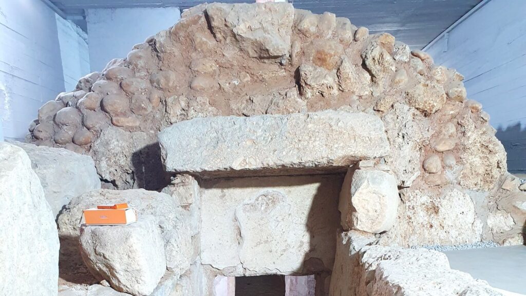 Άποψη από ψηλά του αρχαιολογικού χώρου που δείχνει την πέτρινη καμάρα του Μακεδονικού τάφου και δύο παράλληλους κιβωτιόσχημους τάφους στο προσκήνιο, οι οποίοι οριοθετούν τον διάδρομο πρόσβασης.