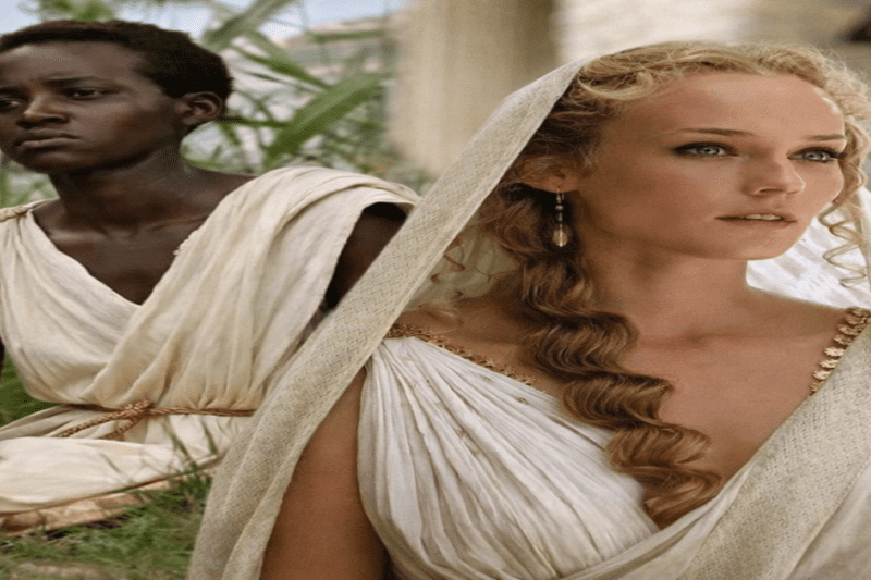 Δύο γυναικείες μορφές με αρχαιοελληνική ενδυμασία, η μία με σκούρα επιδερμίδα και η άλλη με ξανθά μαλλιά και πέπλο, σε υπαίθριο σκηνικό που παραπέμπει στην αρχαιότητα.