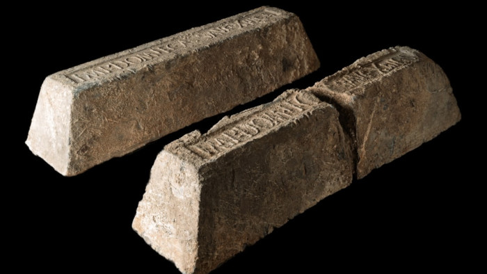 Κοντινό πλάνο δύο αρχαίων ρωμαϊκών ράβδων μολύβδου σε σχήμα τραπεζίου πάνω σε μαύρο φόντο. Φέρουν την επιγραφή "IMP DOMIT AVG..." που παραπέμπει στον Αυτοκράτορα Δομιτιανό.