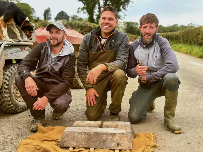Τρεις άνδρες χαμογελούν γονατισμένοι πίσω από τα αρχαιολογικά ευρήματα (τις ράβδους μολύβδου) που είναι τοποθετημένα πάνω σε ένα λινάτσα σε έναν αγροτικό δρόμο.