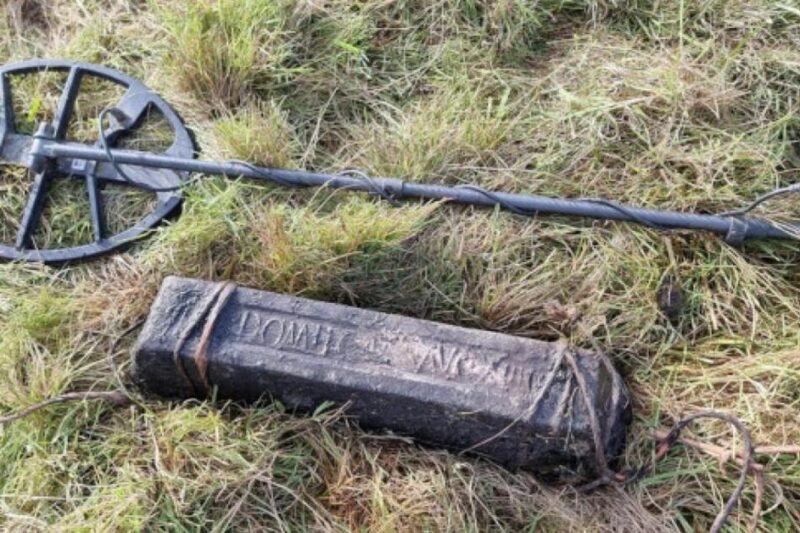 Μια μεγάλη, ορθογώνια ράβδος μολύβδου με ανάγλυφη λατινική επιγραφή, τοποθετημένη στο γρασίδι δίπλα σε έναν σύγχρονο ανιχνευτή μετάλλων.