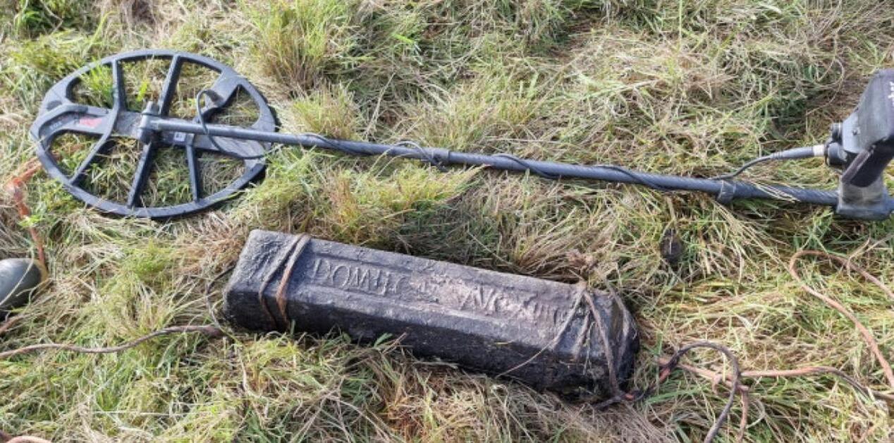 Μια μεγάλη, ορθογώνια ράβδος μολύβδου με ανάγλυφη λατινική επιγραφή, τοποθετημένη στο γρασίδι δίπλα σε έναν σύγχρονο ανιχνευτή μετάλλων.