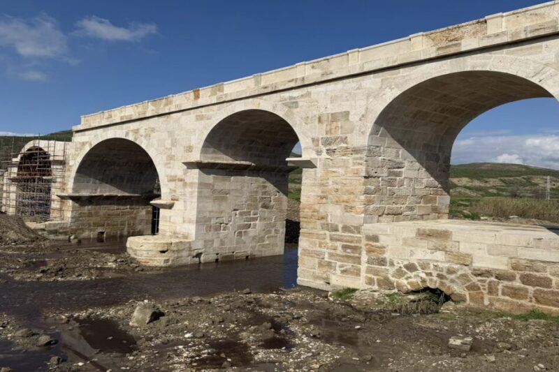 Πλάγια λήψη της ρωμαϊκής γέφυρας του Ασωπού στη Λαοδίκεια, με τρεις ολοκληρωμένες καμάρες από καθαρό λίθο και μια τέταρτη υπό κατασκευή με σκαλωσιές. Νερό ρέει στην κοίτη του ποταμού κάτω από τις καμάρες, σε ανώμαλο έδαφος.