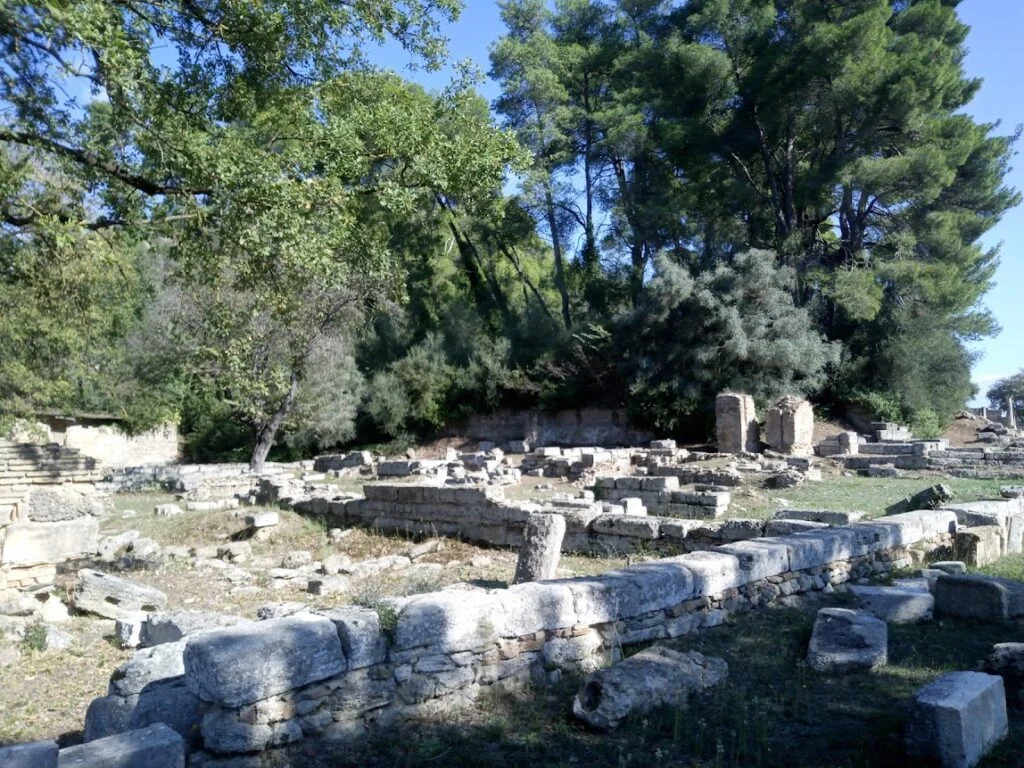 Αρχαιολογικά ερείπια και πέτρινα θεμέλια του Πρυτανείου στην Αρχαία Ολυμπία ανάμεσα σε δέντρα.
