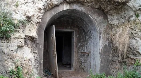 Μια εξωτερική άποψη της εισόδου ενός αντιαεροπορικού καταφυγίου, λαξευμένου μέσα σε έναν βραχώδη λόφο με ξερά χόρτα και θάμνους. Η είσοδος είναι μια τσιμεντένια καμάρα, με μια βαριά, σκουριασμένη μεταλλική πόρτα ανοιχτή. Το εσωτερικό είναι σκοτεινό.