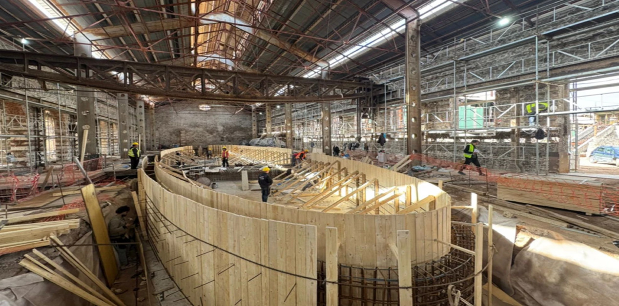 Μια φωτογραφία του εσωτερικού ενός βιομηχανικού κτιρίου στο Τεχνολογικό Πολιτιστικό Πάρκο Λαυρίου, όπου εργάτες με κράνη εργάζονται σε ένα μεγάλο ξύλινο καλούπι σκυροδέματος για τα θεμέλια του υπερυπολογιστή.