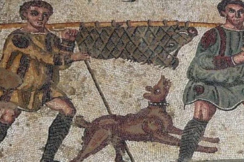 Αρχαίο μωσαϊκό που απεικονίζει δύο κυνηγούς με παραδοσιακή ενδυμασία να μεταφέρουν ένα θήραμα παγιδευμένο σε δίχτυ πάνω σε ένα ξύλινο κοντάρι, ενώ ένας κυνηγετικός σκύλος τους ακολουθεί με ενθουσιασμό.