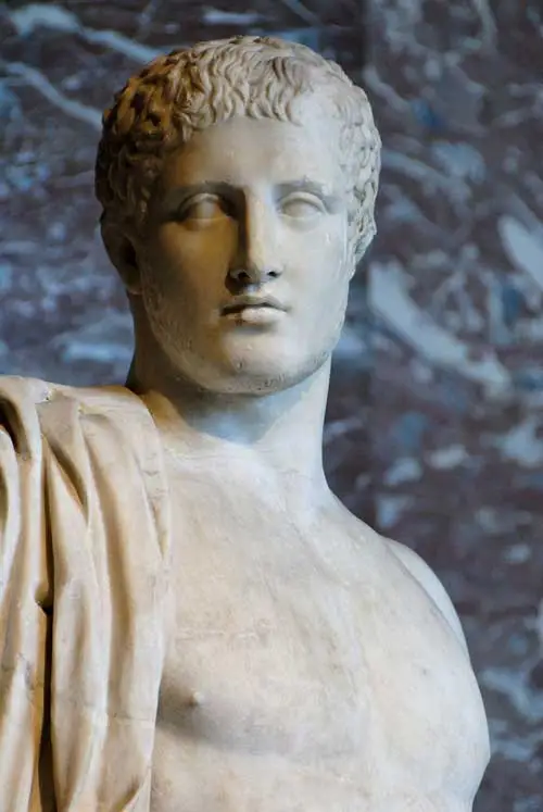 Κοντινό πλάνο σε μαρμάρινο άγαλμα ενός εξιδανικευμένου νεαρού άνδρα με κοντά, σγουρά μαλλιά και έναν μανδύα ριγμένο στον ώμο του.