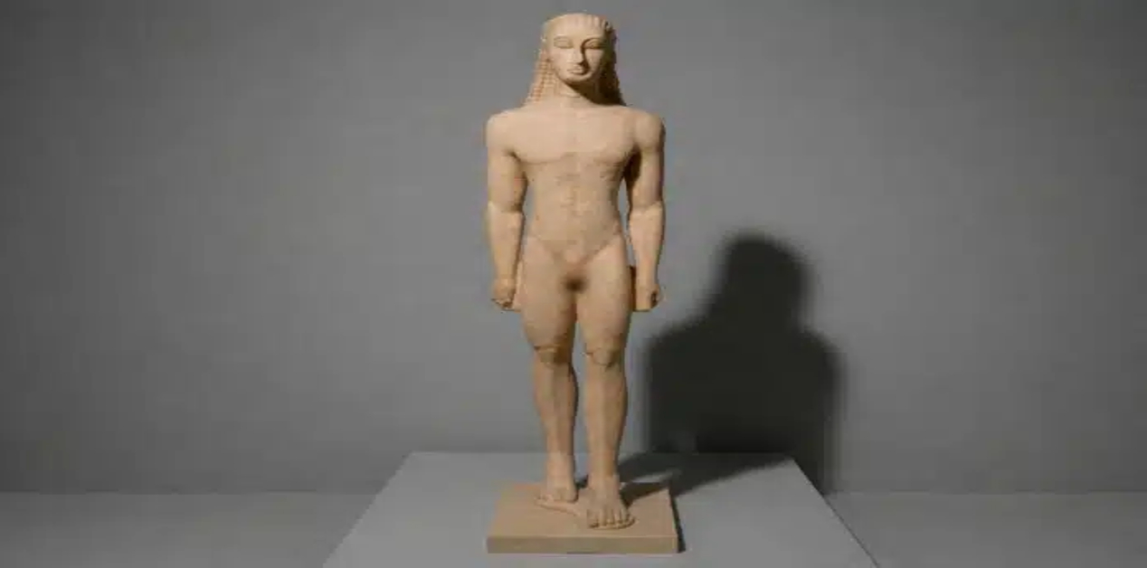Γιατί τα αρχαία ελληνικά αγάλματα είχαν το αριστερό πόδι μπροστά; - Μαρμάρινο άγαλμα ενός γυμνού νεαρού άνδρα (Κούρου) που στέκεται με το αριστερό πόδι μπροστά σε ένα γκρι φόντο.