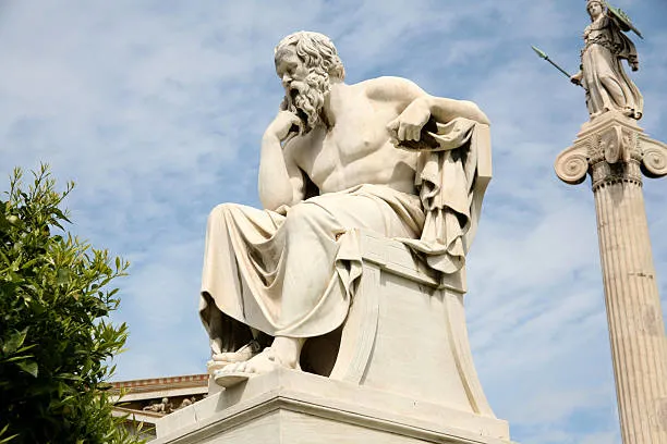 Μαρμάρινο άγαλμα του φιλοσόφου Σωκράτη σε καθιστή στάση συλλογισμού, με το άγαλμα της θεάς Αθηνάς στο φόντο.
