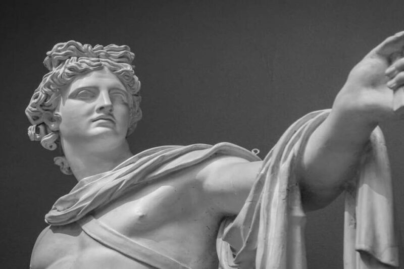 Ασπρόμαυρη φωτογραφία της προτομής και του αριστερού χεριού ενός αρχαίου μαρμάρινου αγάλματος (του Απόλλωνα), το οποίο κοιτάζει στο πλάι και φέρει έναν μανδύα ριγμένο στον βραχίονά του.