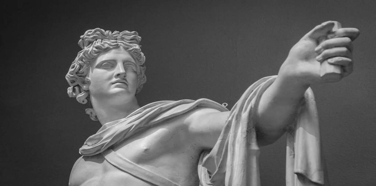 Ασπρόμαυρη φωτογραφία της προτομής και του αριστερού χεριού ενός αρχαίου μαρμάρινου αγάλματος (του Απόλλωνα), το οποίο κοιτάζει στο πλάι και φέρει έναν μανδύα ριγμένο στον βραχίονά του.