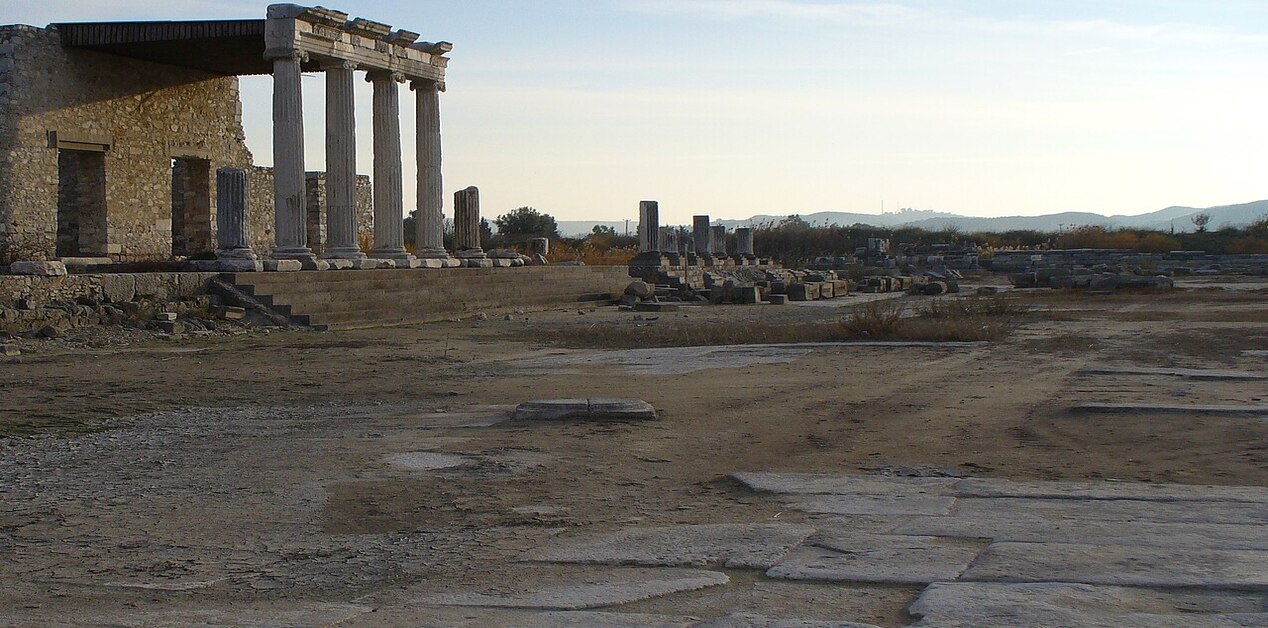 Εκτεταμένος αρχαιολογικός χώρος της Μιλήτου στην Ιωνία της Μικράς Ασίας, που δείχνει τα ερείπια μιας δωρικής κιονοστοιχίας, ανακατασκευασμένες κατασκευές και εκτεταμένα θεμέλια κάτω από ένα απαλό απογευματινό φως.