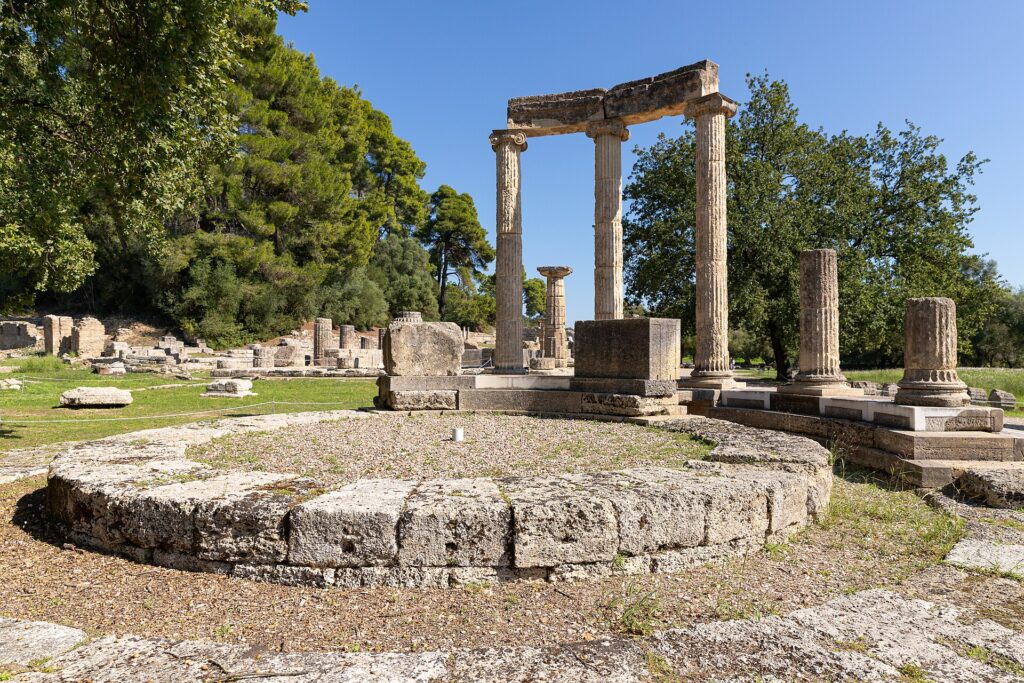 Έγχρωμη φωτογραφία που δείχνει τα ερείπια του Ναού της Ήρας στην Ολυμπία, με τρεις σωζόμενους δωρικούς κίονες που στηρίζουν μέρος του επιστυλίου, περιτριγυρισμένους από πεύκα και ελαιόδεντρα, κάτω από γαλάζιο ουρανό.