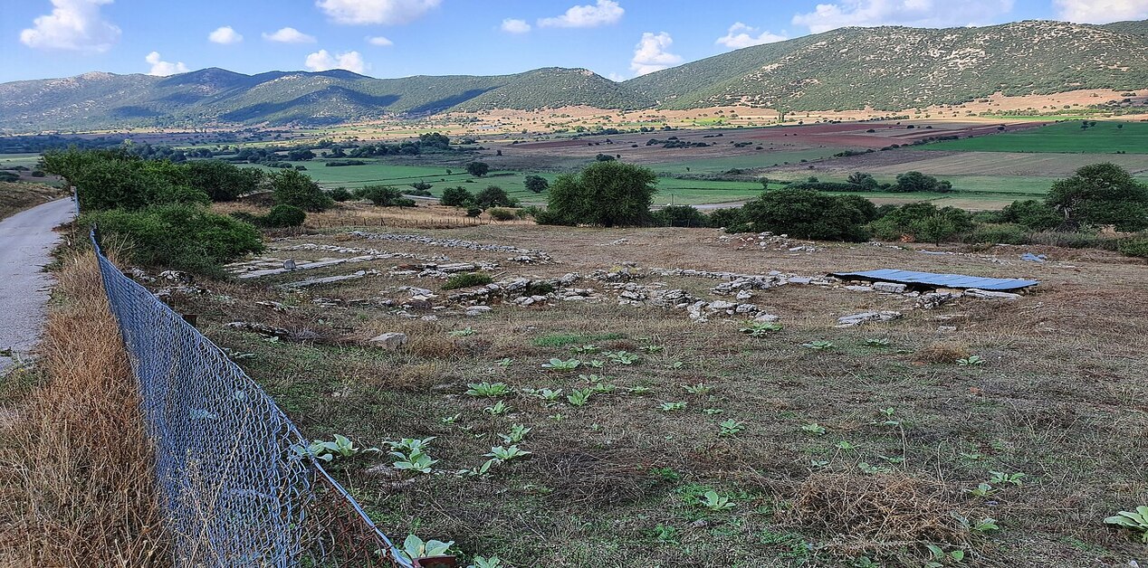 Αρχαιολογικός χώρος με πέτρινα θεμέλια αρχαίων κτισμάτων σε μια καταπράσινη κοιλάδα, περιτριγυρισμένος από βουνά κάτω από έναν γαλάζιο ουρανό με σύννεφα.