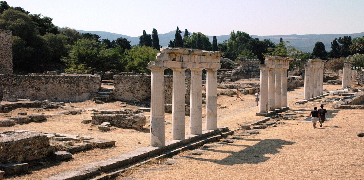 Μια σειρά από αρχαίους ελληνικούς κίονες που στέκονται ανάμεσα σε πέτρινα ερείπια στην Αρχαία Αγορά της Κω, με δέντρα και λόφους στο βάθος υπό καθαρό ουρανό. Τουρίστες περπατούν ανάμεσα στα ερείπια