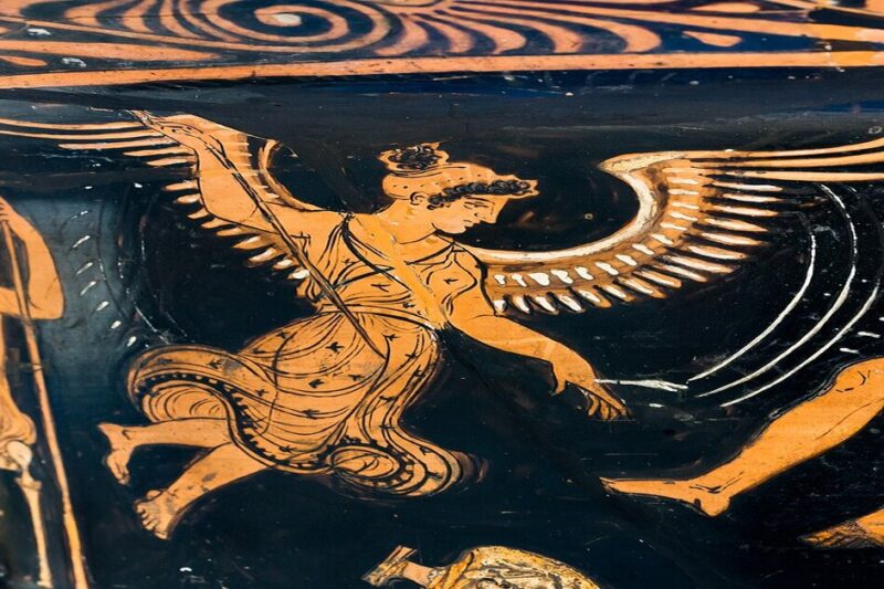 Κοντινή φωτογραφία τμήματος αρχαίου ελληνικού ερυθρόμορφου αγγείου. Μια πτερωτή γυναικεία μορφή σε κίνηση, με περίτεχνα φτερά και φόρεμα, περιβάλλεται από άλλες μορφές.
