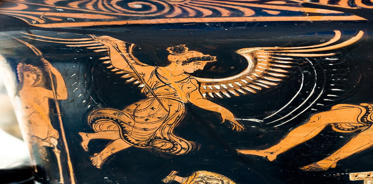 Κοντινή φωτογραφία τμήματος αρχαίου ελληνικού ερυθρόμορφου αγγείου. Μια πτερωτή γυναικεία μορφή σε κίνηση, με περίτεχνα φτερά και φόρεμα, περιβάλλεται από άλλες μορφές.