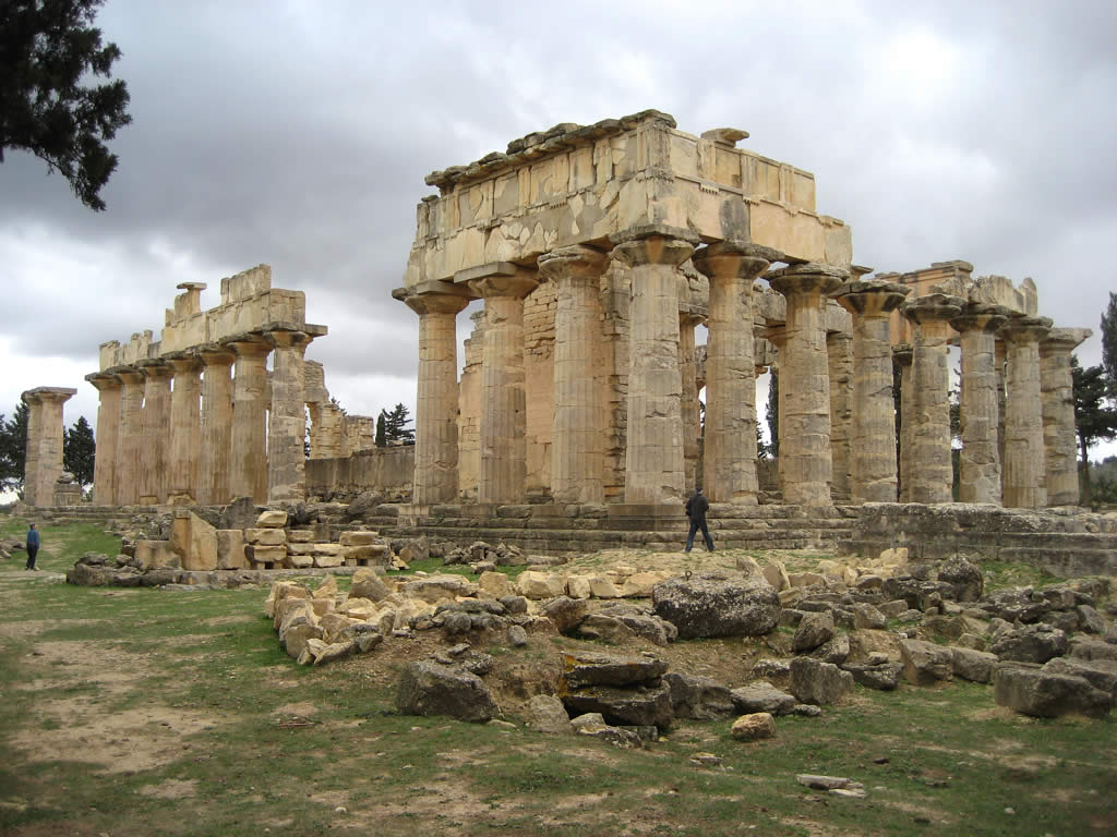 Φωτογραφία των ερειπίων ενός μεγάλου αρχαίου ελληνικού δωρικού ναού στην Κυρήνη, Λιβύη, με πολλούς όρθιους κίονες που υποστηρίζουν τμήματα του επιστυλίου. Πεσμένοι λίθοι περιβάλλουν τη βάση.