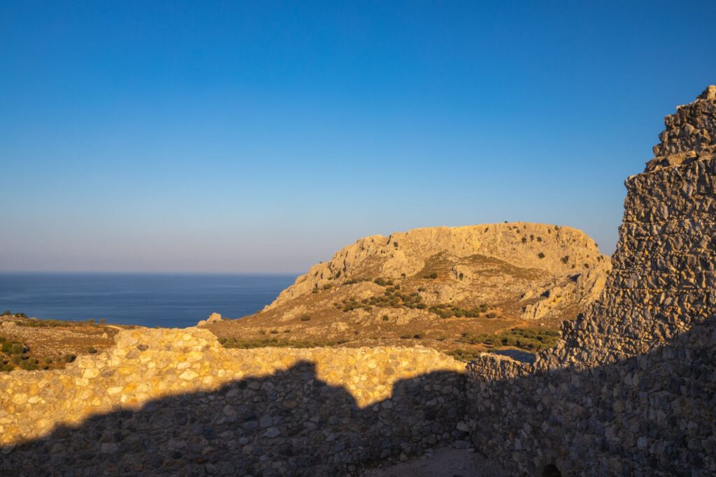 Αλία της Ρόδου: Η Τραγική Θεότητα και το Σκοτεινό Μυστικό του Νησιού 7 Αρχαία πέτρινα τείχη με θέα έναν βραχώδη λόφο και την καταγάλανη θάλασσα της Ρόδου.