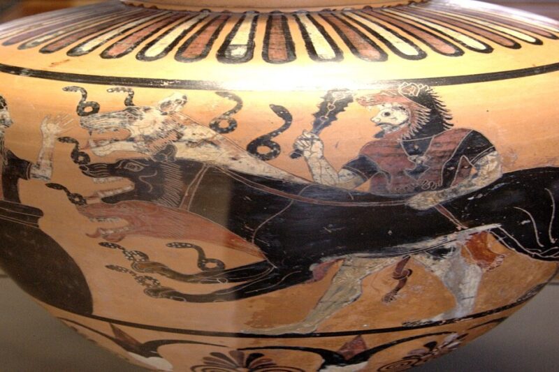 Κοντινό πλάνο ενός αρχαίου ελληνικού μελανόμορφου αγγείου, που απεικονίζει τον Ηρακλή, φορώντας τη λεοντή, να σέρνει με λουρί τον δικέφαλο Κέρβερο, ο οποίος είναι γεμάτος coiling φίδια γύρω από τα κεφάλια του. Αριστερά, μια έντρομη ανδρική φιγούρα (πιθανόν ο Ευρυσθέας) παρακολουθεί μέσα από ένα πιθάρι.