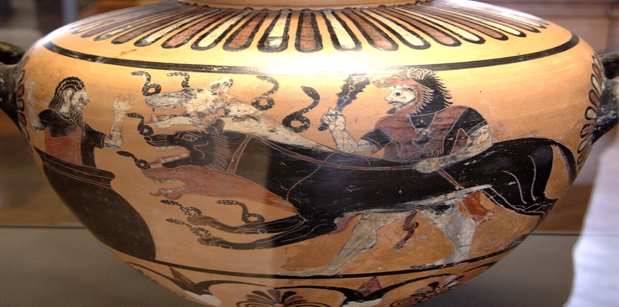 Κοντινό πλάνο ενός αρχαίου ελληνικού μελανόμορφου αγγείου, που απεικονίζει τον Ηρακλή, φορώντας τη λεοντή, να σέρνει με λουρί τον δικέφαλο Κέρβερο, ο οποίος είναι γεμάτος coiling φίδια γύρω από τα κεφάλια του. Αριστερά, μια έντρομη ανδρική φιγούρα (πιθανόν ο Ευρυσθέας) παρακολουθεί μέσα από ένα πιθάρι.
