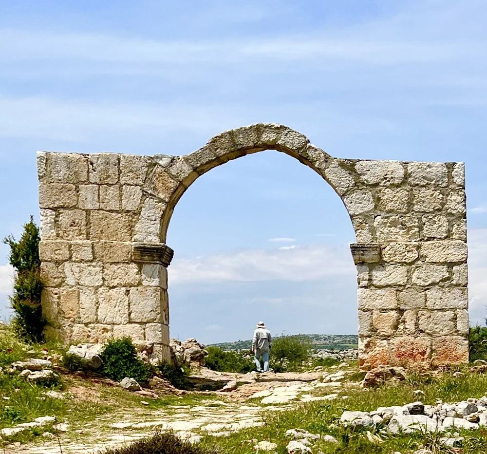 Μια μνημειώδης πέτρινη αψίδα από αρχαία ερείπια στη μέση ενός καταπράσινου λιβαδιού, με έναν άνδρα να περπατά στο βάθος.