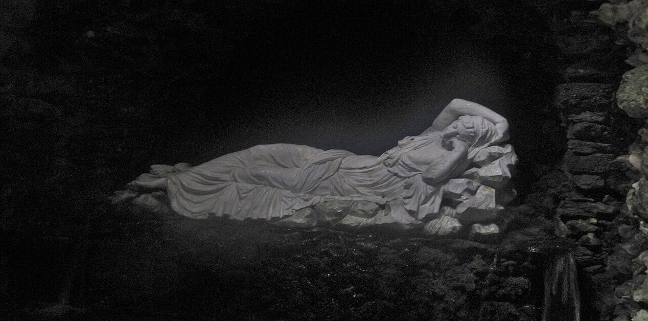 Ένα κλασικό άγαλμα μιας νύμφης που ξαπλώνει ανάμεσα σε βράχους, φωτισμένο από ένα σημείο σε ένα σκοτεινό, σαν σπηλιά περιβάλλον. Η νύμφη Κλεοδώρα (nymfi-kleodora-parnassos.jpg) είναι ντυμένη με πτυχωτό χιτώνα και αναπαύεται, στηριζόμενη στον αγκώνα της.