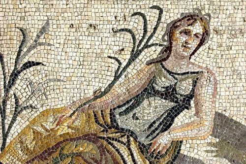 Αρχαίο ψηφιδωτό που δείχνει μια γυναικεία μορφή καθισμένη δίπλα σε φυτά που μοιάζουν με καλαμιές.