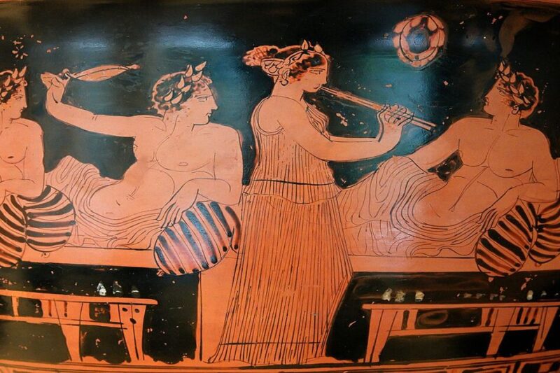 Αρχαία ελληνική ερυθρόμορφη παράσταση συμποσίου σε αγγείο. Τέσσερις άνδρες ανακλίνονται σε κλίνες κρατώντας αντικείμενα ψυχαγωγίας, ενώ μια γυναίκα μουσικός στο κέντρο παίζει διπλό αυλό, δημιουργώντας ατμόσφαιρα γιορτής και διασκέδασης.